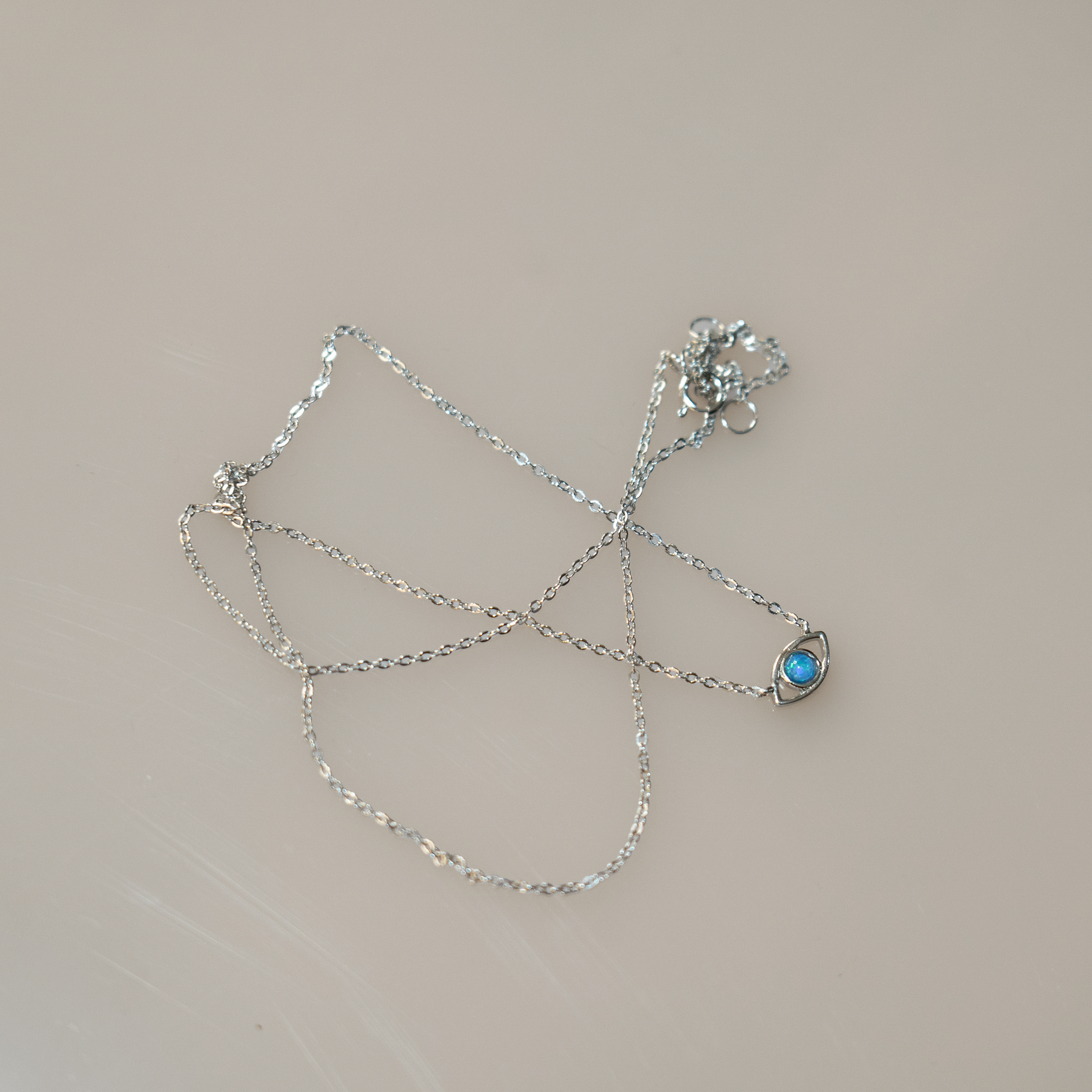 Sterling Silver chain with lobster clasp holds an evil eye charm. The eye is a round blue opal set in gold with a hanging sterling silver chain below. 