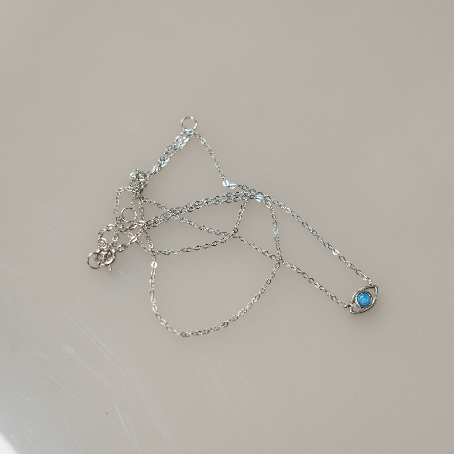 Sterling Silver chain with lobster clasp holds an evil eye charm. The eye is a round blue opal set in gold with a hanging sterling silver chain below. 