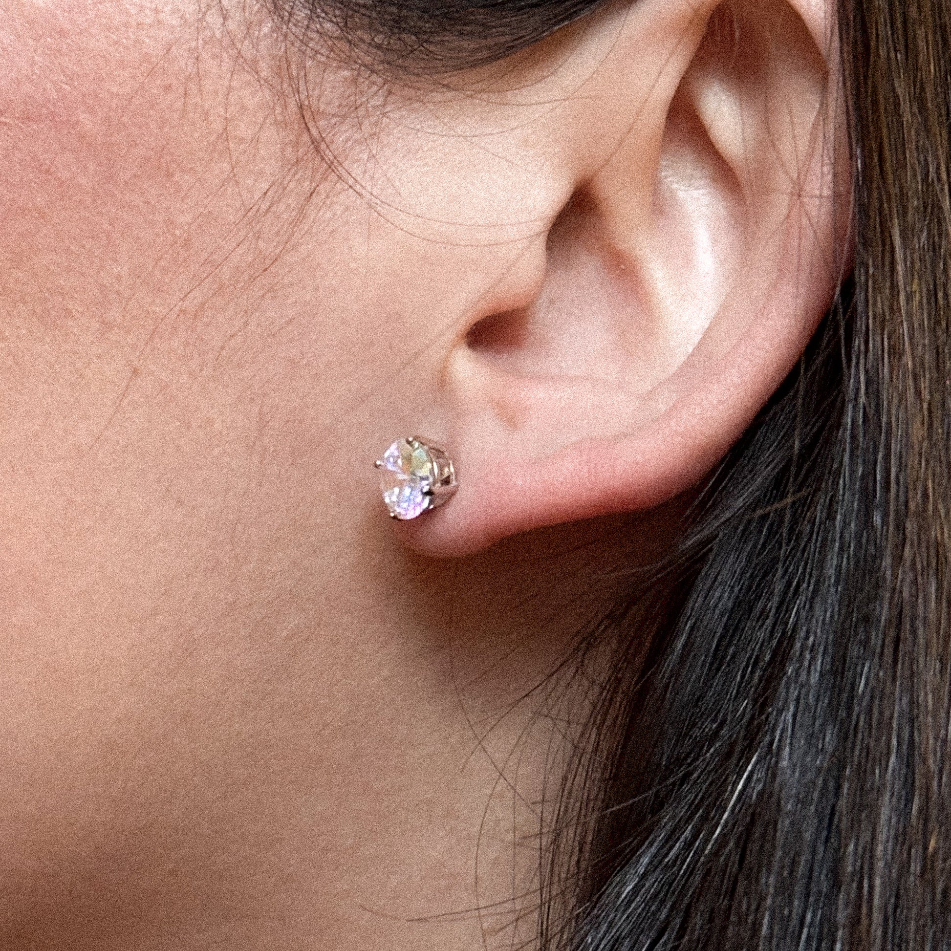 Close-up of an ear wearing a small oval-shaped clear crystal stud earring set in silver. The earring catches the light, appearing bright against the wearers skin and dark hair.