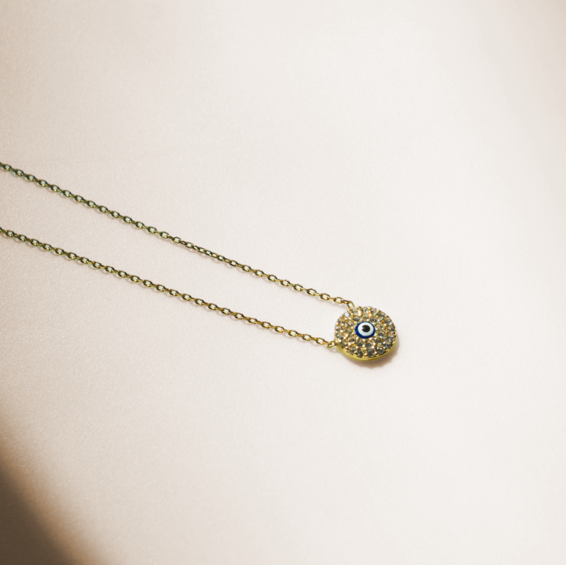 Dainty necklace with a thin gold chain and a small circular pendant. The pendant has a flat disc shape with a round blue cubic zirconia stone in the center surrounded by smaller clear stones, forming an eye design. Gold in color.