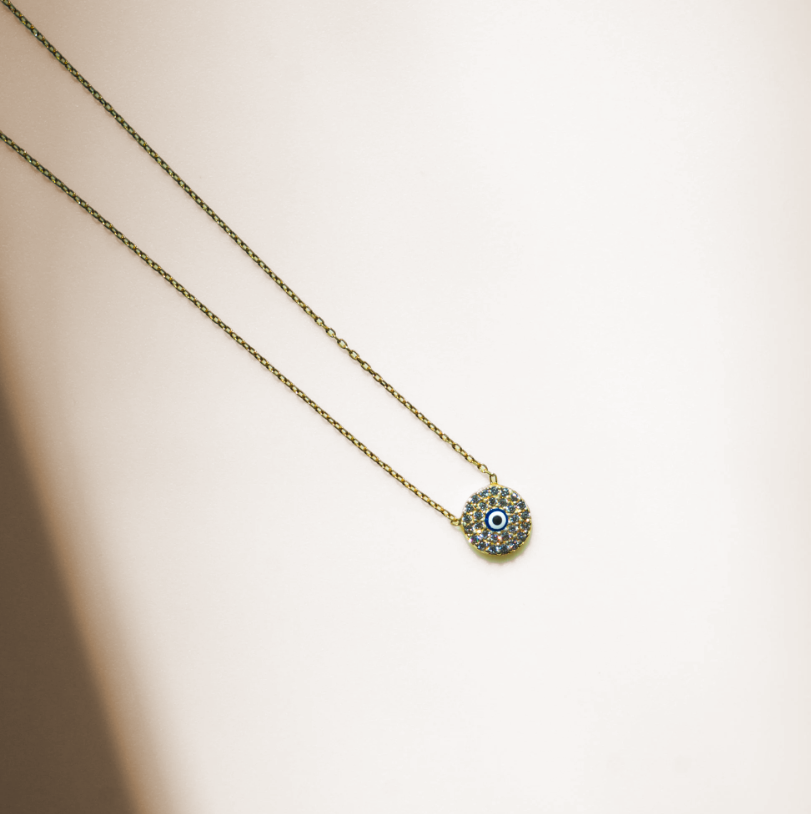 Dainty necklace with a thin gold chain and a small circular pendant. The pendant has a flat disc shape with a round blue cubic zirconia stone in the center surrounded by smaller clear stones, forming an eye design. Gold in color.