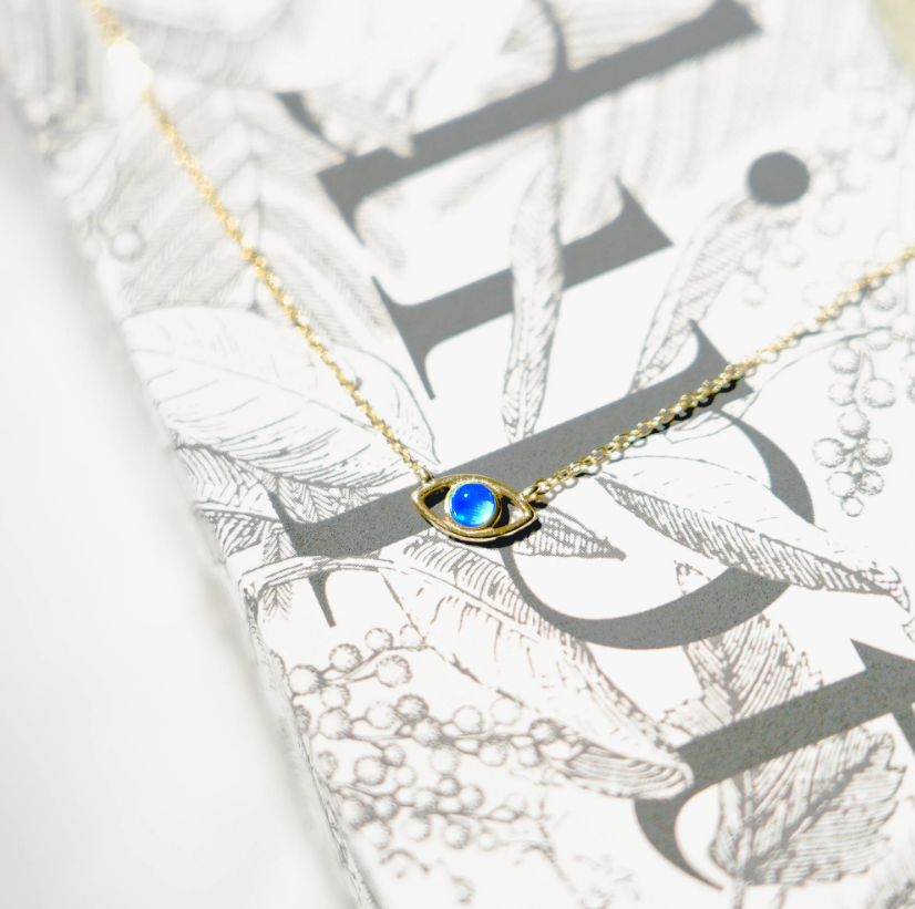 Gold vermeil chain with lobster clasp holds an evil eye charm. The eye is a round blue opal set in gold with a hanging gold vermeil chain below. Sterling silver and opal, gold-plated.