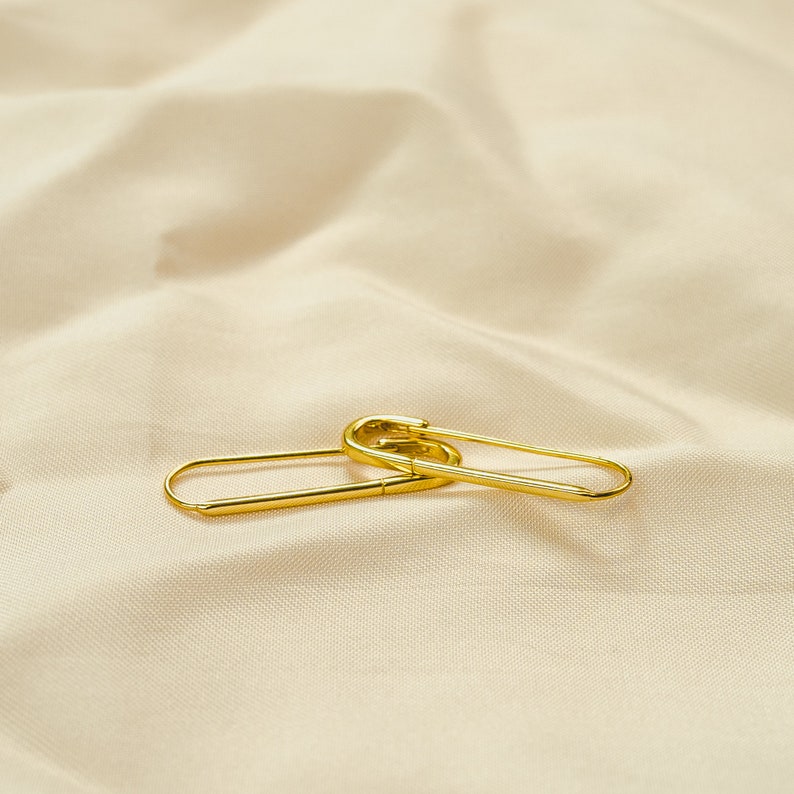 These earrings feature an elongated paperclip-inspired design crafted in gleaming 18k gold vermeil. The smooth, curved shape forms an open hoop that hooks directly into the earlobe. The minimalist style complements the warm gold tones beautifully.