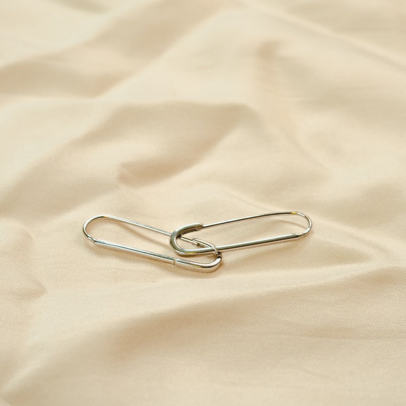 These earrings feature an elongated paperclip-inspired design crafted in gleaming 925 sterling silver. The smooth, curved shape forms an open hoop that hooks directly into the earlobe. The minimalist style complements the warm gold tones beautifully.