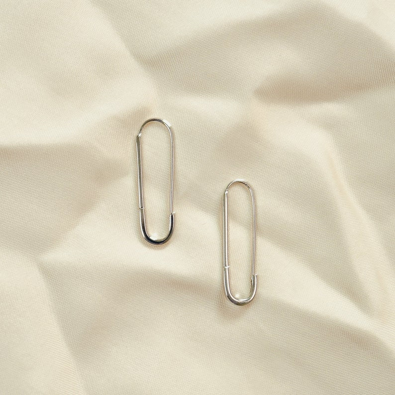 These earrings feature an elongated paperclip-inspired design crafted in gleaming 925 sterling silver. The smooth, curved shape forms an open hoop that hooks directly into the earlobe. The minimalist style complements the warm gold tones beautifully.