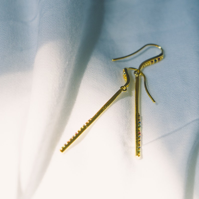 These dangle earrings feature a sleek gold vermeil bar adorned with a row of colorful cubic zirconia stones in a rainbow pattern. They have a fish hook closure for easy wear. The vibrant gems complement the warm gold tones beautifully.