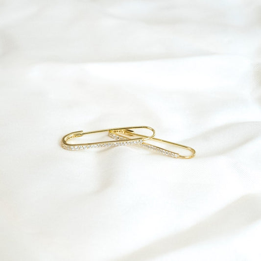 These earrings feature an elongated paperclip-inspired design in gold vermeil with a row of small cubic zirconia stones along one side. The smooth, curved shape forms an open hoop that hooks directly into the earlobe, combining minimalism with subtle sparkle.