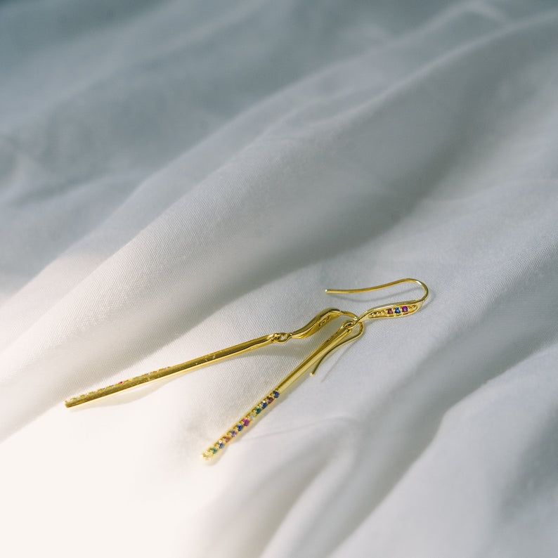 These dangle earrings feature a sleek gold vermeil bar adorned with a row of colorful cubic zirconia stones in a rainbow pattern. They have a fish hook closure for easy wear. The vibrant gems complement the warm gold tones beautifully.