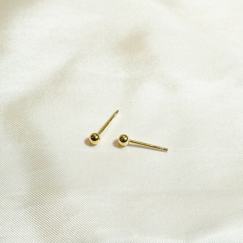 Gold ball stud earrings on thin post. Polished round beads, post and butterfly closure. 2mm diameter. Solid 14K gold, no plating or gemstones. Minimalist style.