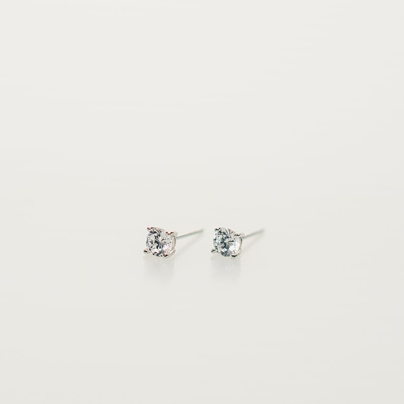 These earrings feature small, square-cut clear gemstone cubic zirconia, set in sleek sterling silver studs. The minimalist design complements various ear piercings, from cartilage to lobes.