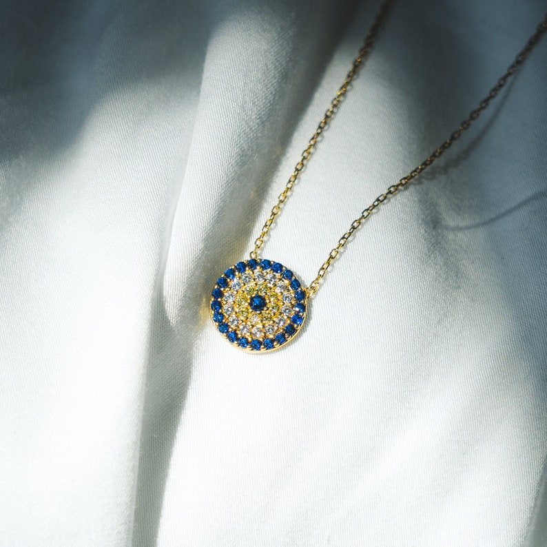 Round evil eye pendant necklace, 12mm diameter, crafted in 18k gold vermeil (thick gold layer over sterling silver). Blue and white cubic zirconia stones set in pendant. Adjustable chain length from 15 to 17 inches. Lobster claw clasp closure.