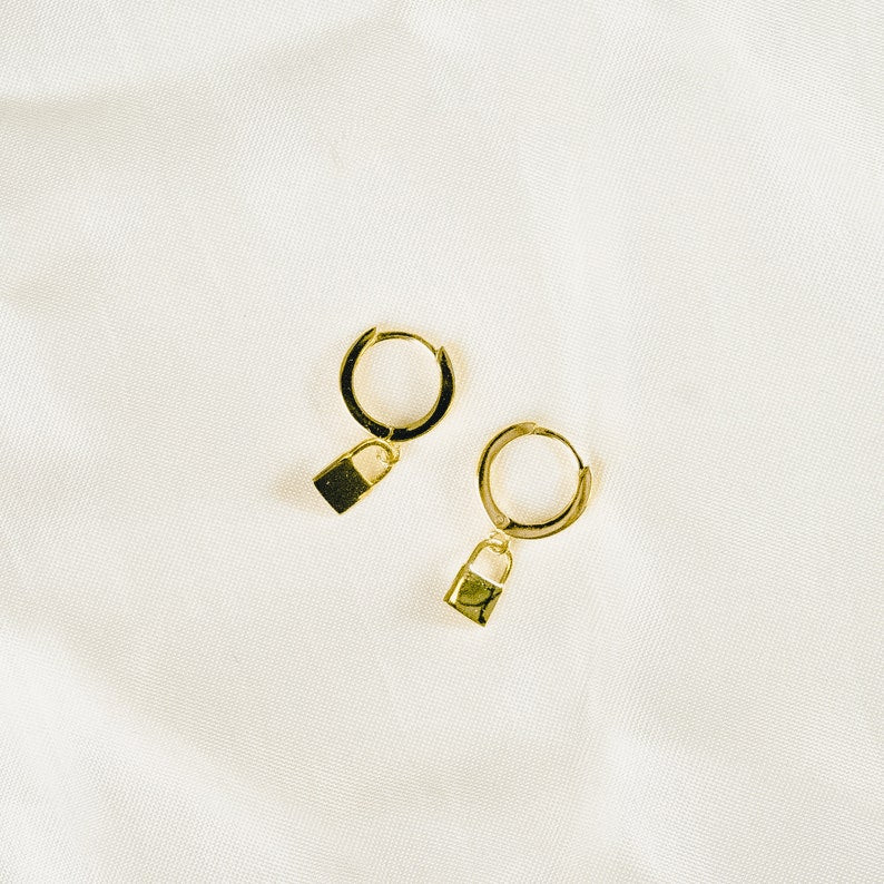 These earrings feature small gold vermeil hoop earrings with tiny dangling padlock charms. The simple, minimalist design showcases the warm golden color against a light background.