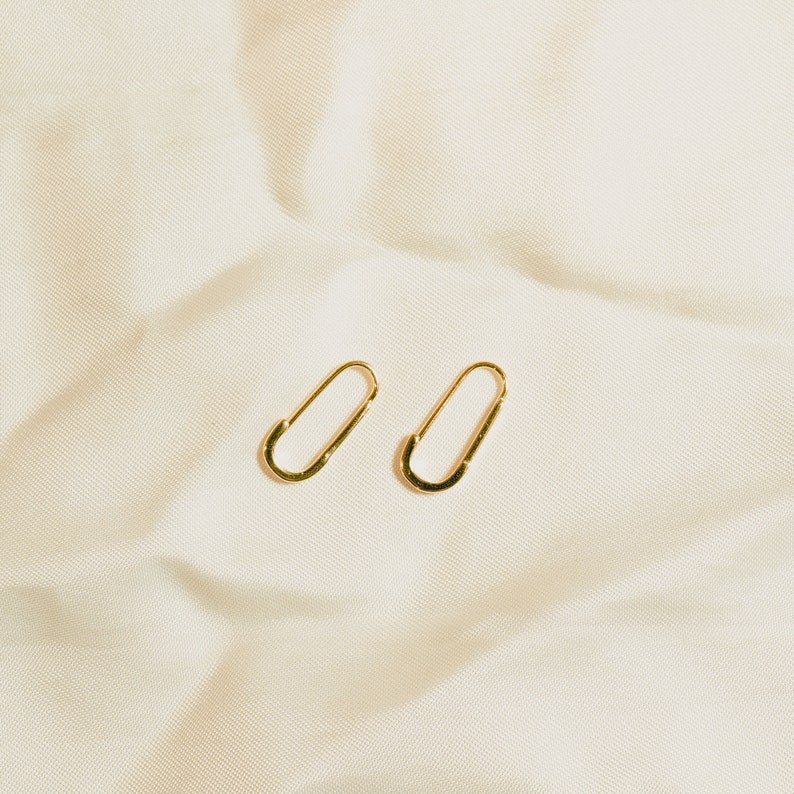 These earrings feature a simple, elongated paperclip design in gleaming 18k gold vermeil. The smooth, curved shape forms a delicate open hoop that hooks directly into the earlobe, embodying minimalist chic.