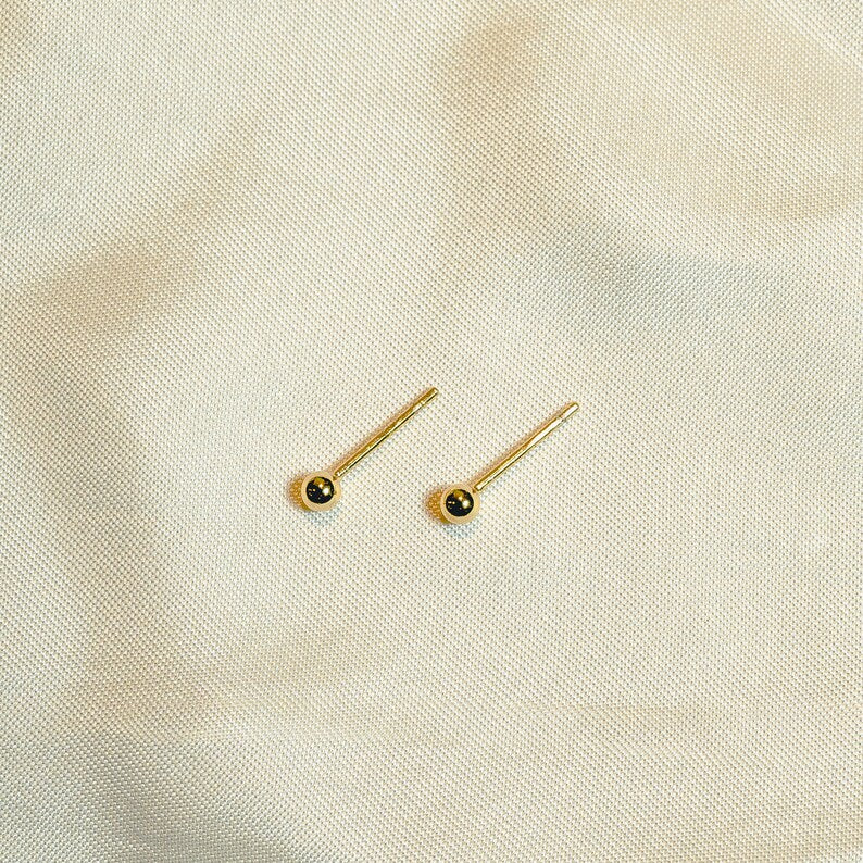 Gold ball stud earrings on thin post. Polished round beads, post and butterfly closure. 2mm diameter. Solid 14K gold, no plating or gemstones. Minimalist style.