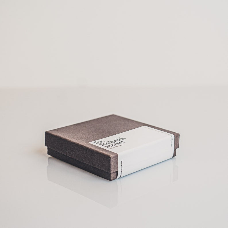 The photo shows the Bushwick Markets packaging a minimalist packaging for shipping items, including a charcoal gray box and a square card in black and white tones, conveying the brands boho aesthetic.