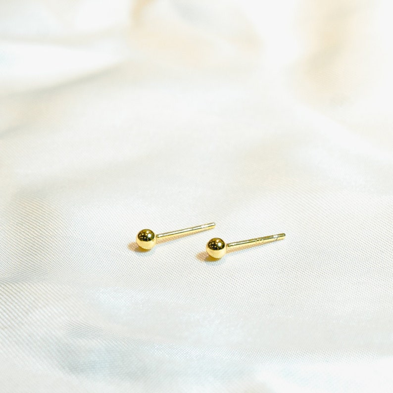 Gold ball stud earrings on thin post. Polished round beads, post and butterfly closure. 2mm diameter. Solid 14K gold, no plating or gemstones. Minimalist style.