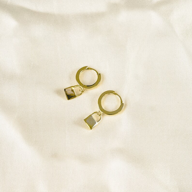 These earrings feature small gold vermeil hoop earrings with tiny dangling padlock charms. The simple, minimalist design showcases the warm golden color against a light background.